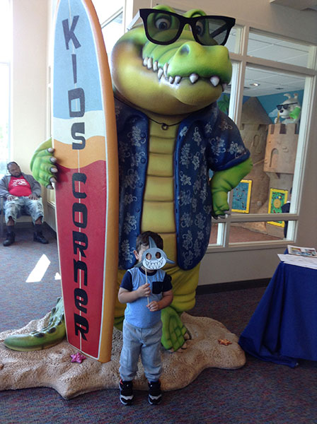 Life-size alligator statue in sunglasses holding a Kids Korner surfboard